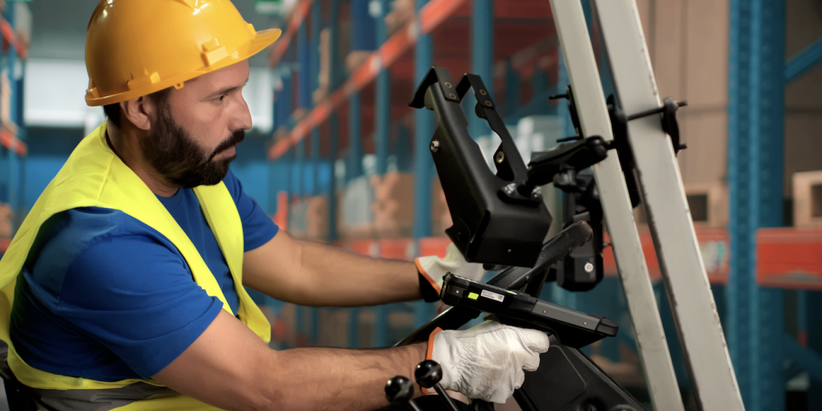 A warehouse worker wearing a yellow hard hat, safety vest, and gloves operates a forklift in a storage area with shelves filled with boxes, efficiently managing inventory using an ERP system.