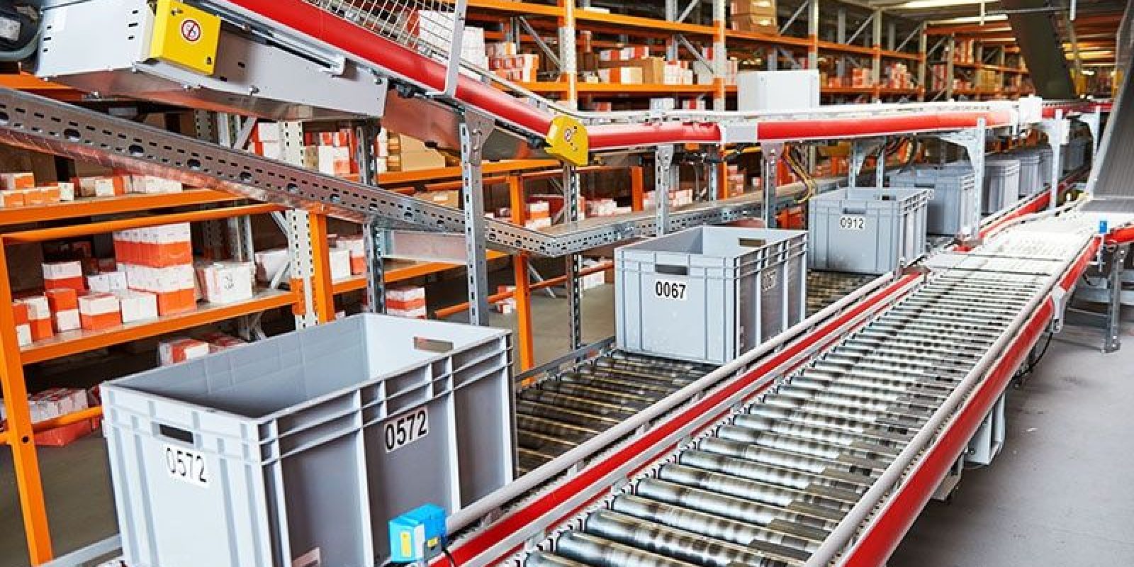 Plastic bins labeled with numbers move along a conveyor belt in a warehouse filled with shelves of orange and white packages. The setting appears organized and industrial, suggesting a logistics or distribution center.