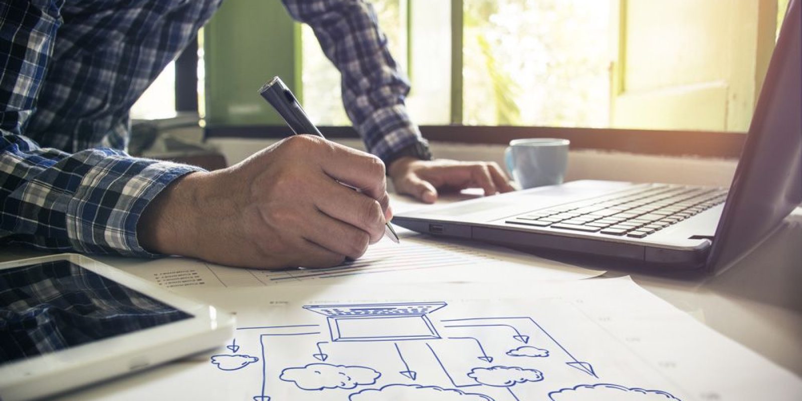 A person in a plaid shirt writes on paper next to a laptop, tablet, and a cloud computing diagram featuring D365 Business Central, with sunlight streaming through a window in the background.