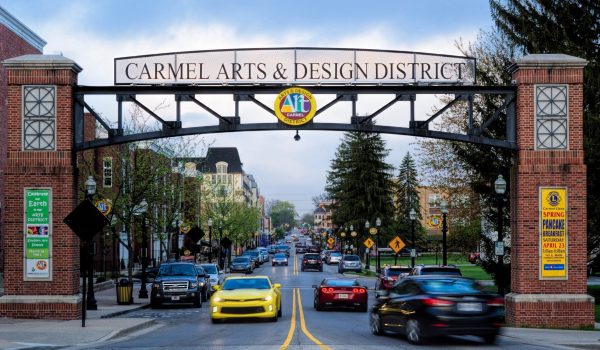 A street scene in Carmel Arts & Design District, Indiana, with a large archway sign overhead, cars driving on the road, trees, buildings, and people walking on the sidewalks. A yellow car is prominent in the foreground.