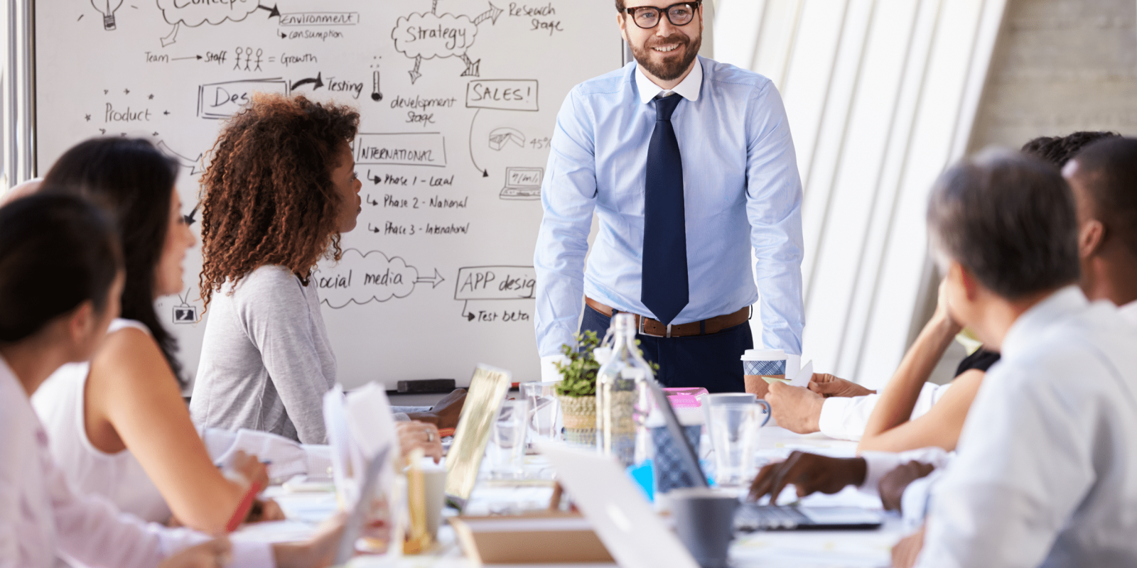 A man in business attire stands and smiles while presenting to a group of colleagues seated around a conference table, with a whiteboard full of notes and diagrams visible in the background.