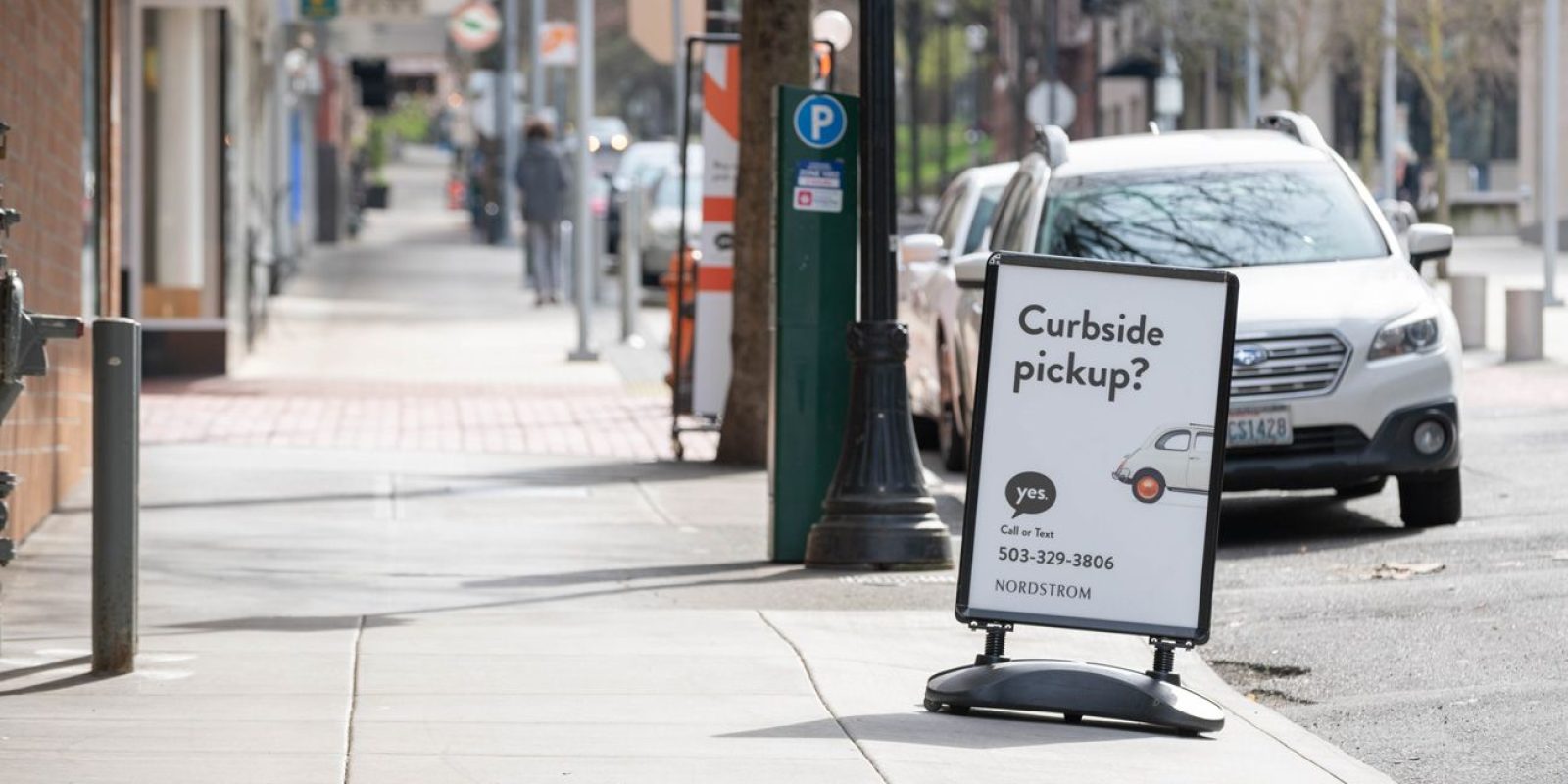A sidewalk sign reads “Curbside pickup? yes” with a phone number and a small car graphic, promoting Nordstrom’s curbside service. Cars are parked on the street and pedestrians walk in the background.