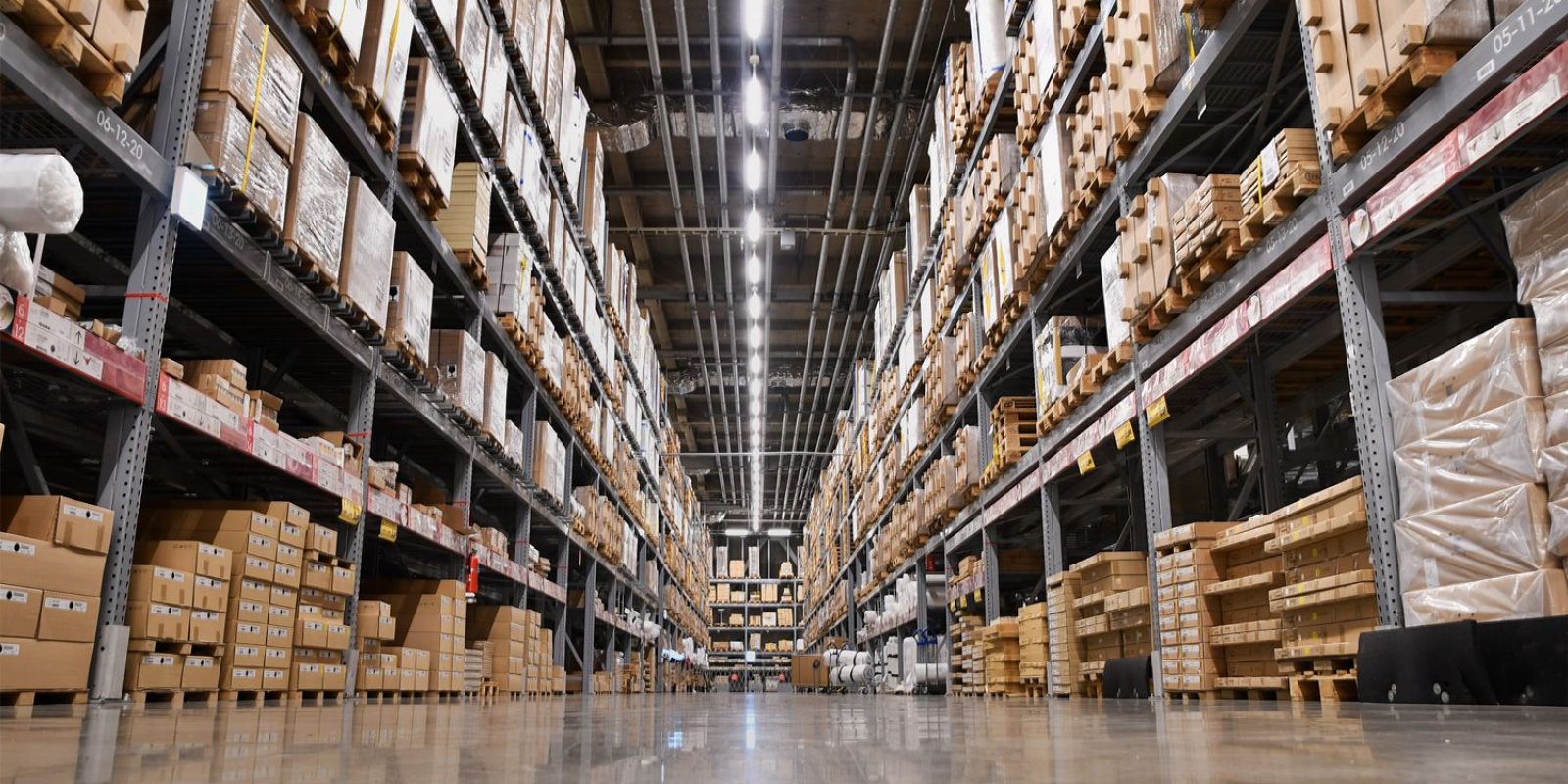 View of a large warehouse with tall shelves stacked with cardboard boxes, ideal for warehouse capacity analysis, bright overhead lighting, and a shiny concrete floor, seen from a low angle between the aisles.