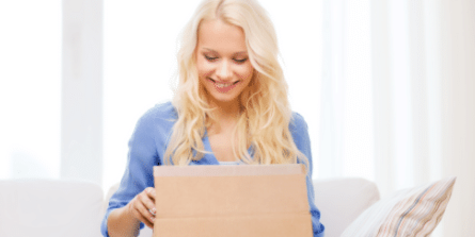 A smiling woman with long blonde hair sits on a couch and opens a cardboard box, looking inside with excitement. A striped pillow is beside her, and soft daylight fills the room.