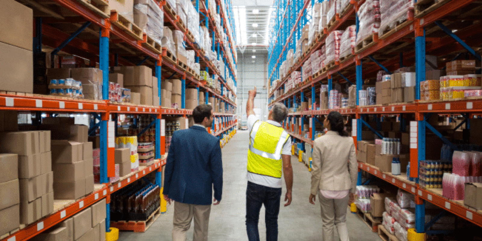 Three people walk down an aisle in a large warehouse, surrounded by tall shelves stocked with boxes and products. One person wears a yellow safety vest, while the others are in business attire.