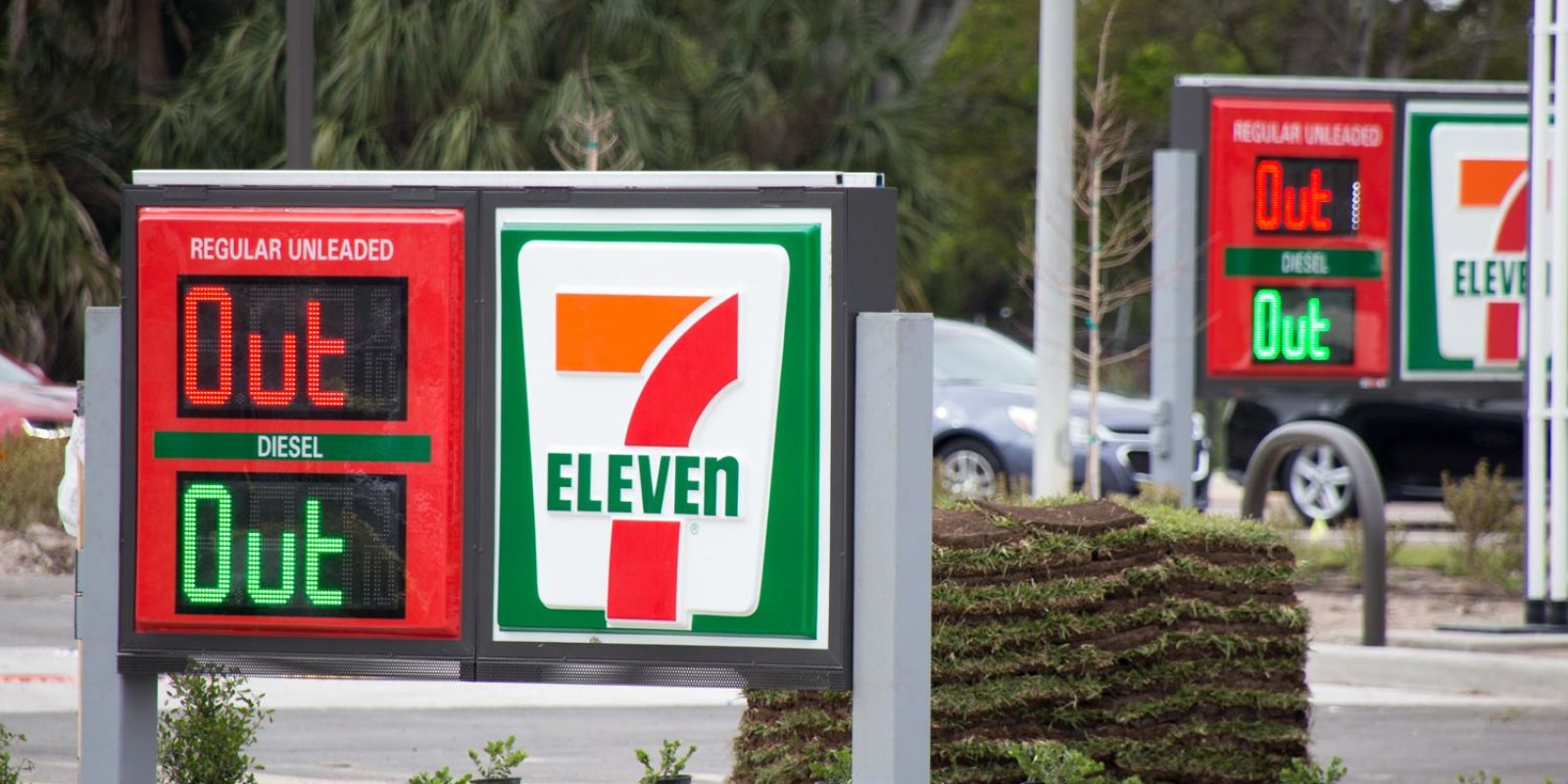 A 7-Eleven gas station sign displays OUT for both regular unleaded and diesel fuel, indicating they are unavailable. The same message appears on a second sign in the background. Small plants line the area.
