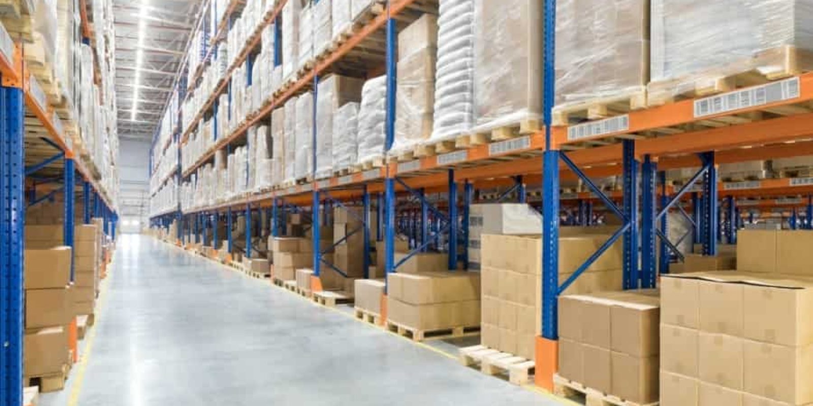Large warehouse interior with tall blue and orange industrial shelving, stacked with cardboard boxes and wrapped pallets; the polished concrete floor and wide aisle highlight a space designed for efficient warehouse automation.