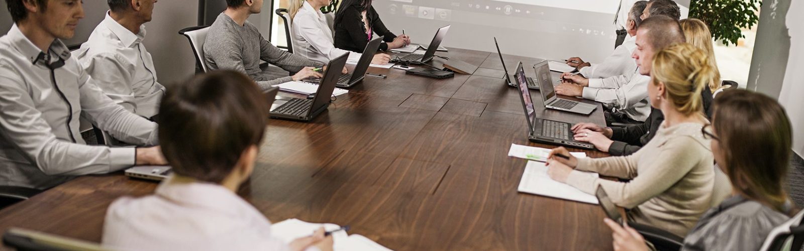 A group of people in business attire sit around a conference table with laptops and notepads, watching a presenter point to a bar chart on a projected screen during a meeting about the Microsoft Dynamics AX upgrade.