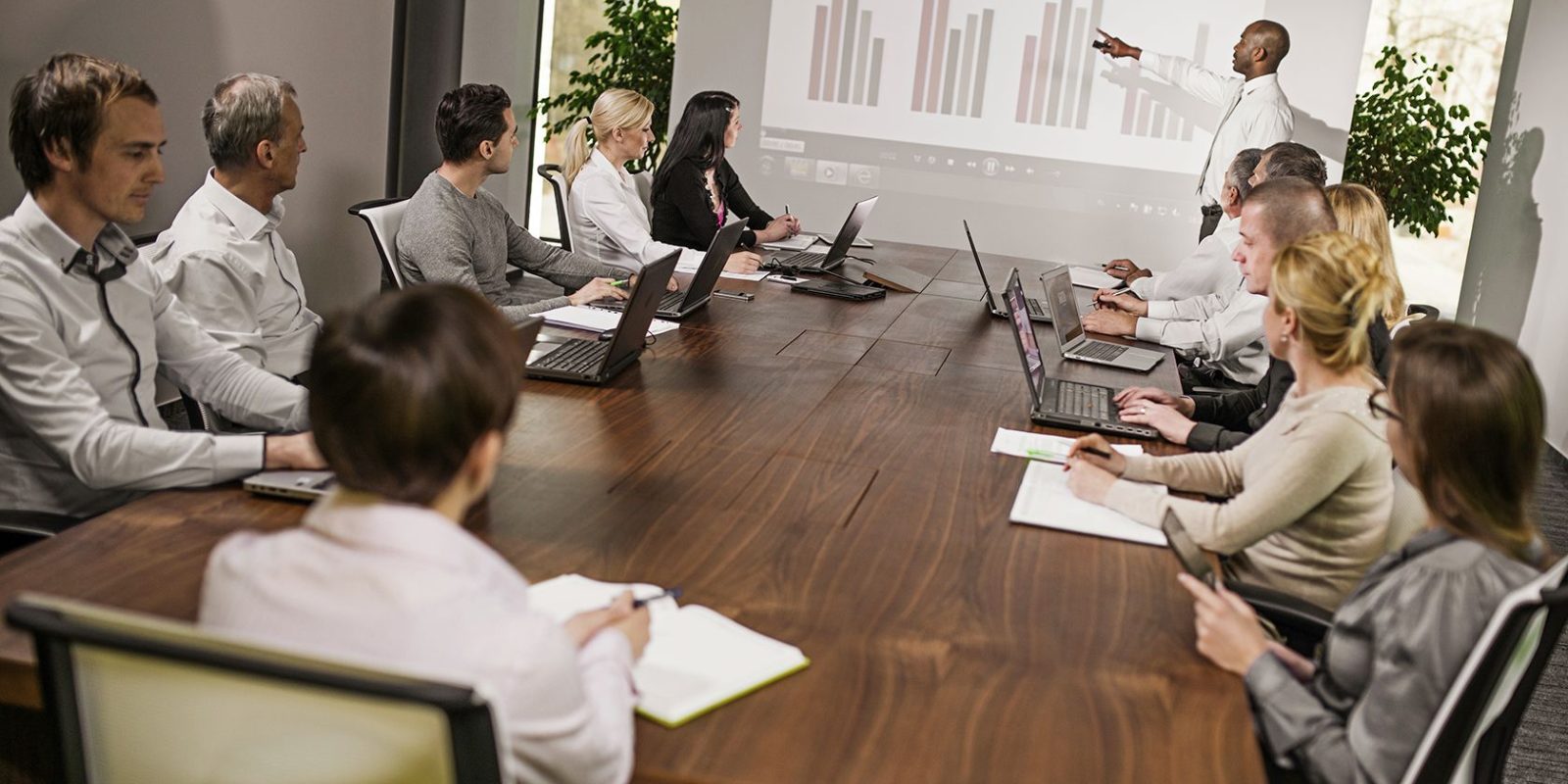 A group of people in business attire sit around a conference table with laptops and notepads, watching a presenter point to a bar chart on a projected screen during a meeting about the Microsoft Dynamics AX upgrade.