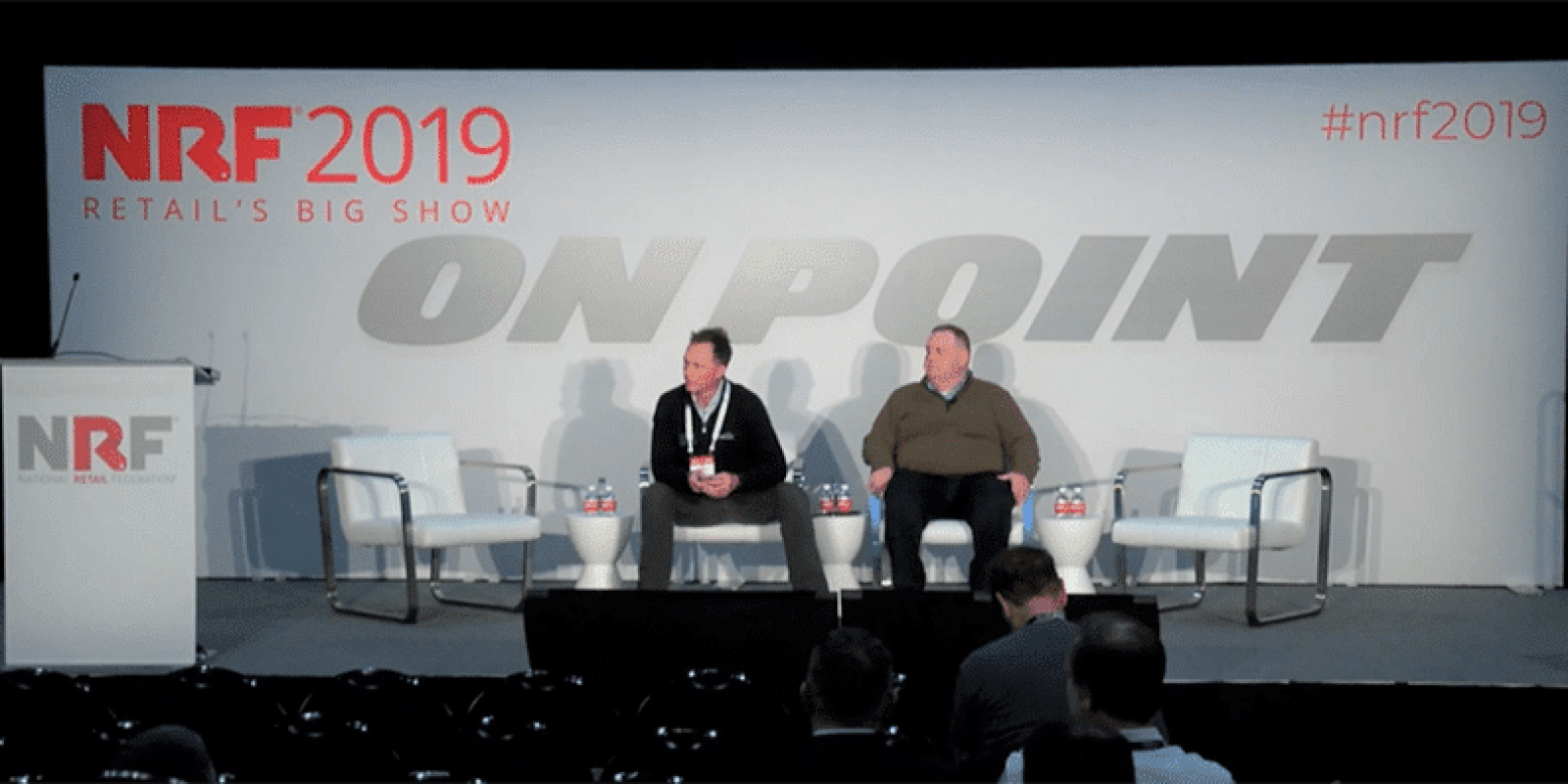 Two men sit on a stage with three empty chairs at NRF 2019 Retail’s Big Show. The backdrop has “ON POINT” and event branding. A podium is on the left, and a few people are seated in the audience.
