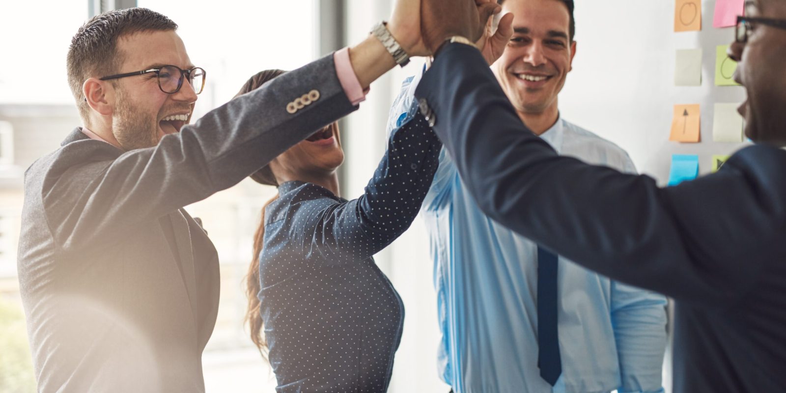 Four business professionals, three men and one woman, smiling and giving a group high-five in an office setting with sticky notes on the wall in the background.