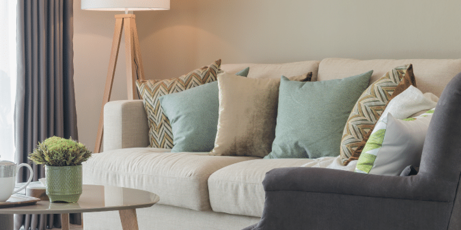 A cozy living room with a beige sofa decorated with green and patterned pillows, a dark gray armchair, a wooden coffee table with a small plant and mugs, a standing lamp, and floor-length curtains.