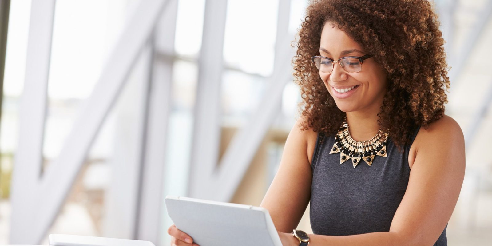 A woman with curly hair and glasses sits at a desk, smiling while looking at a tablet. Papers, a notebook, and a laptop with power platform dashboards open are on the desk in front of her. The background shows a bright, modern interior.