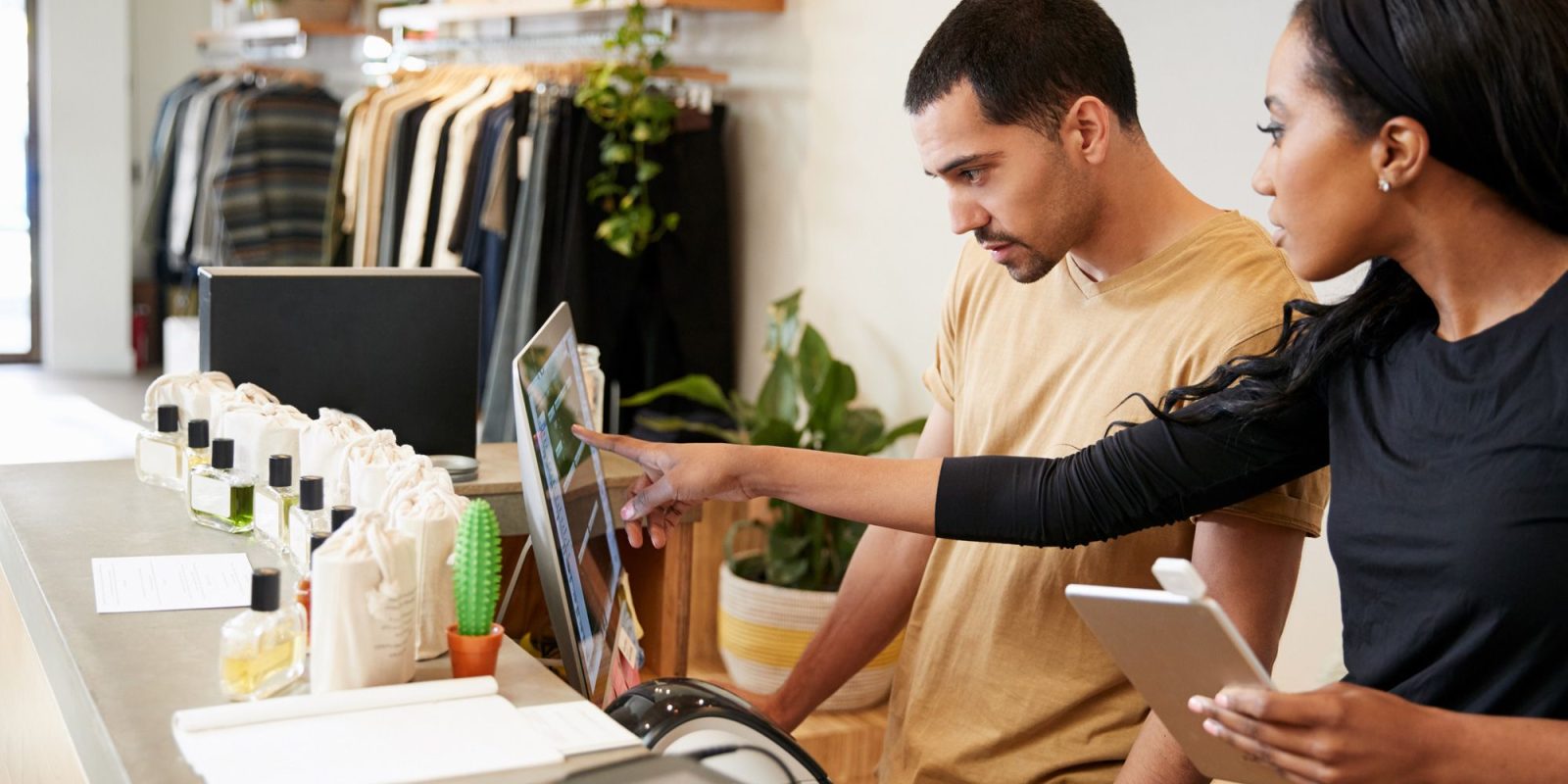 Two people stand at a retail store counter, one pointing at a computer screen while the other holds a tablet—demonstrating retail management systems. Shelves with clothing and various products are visible in the background.
