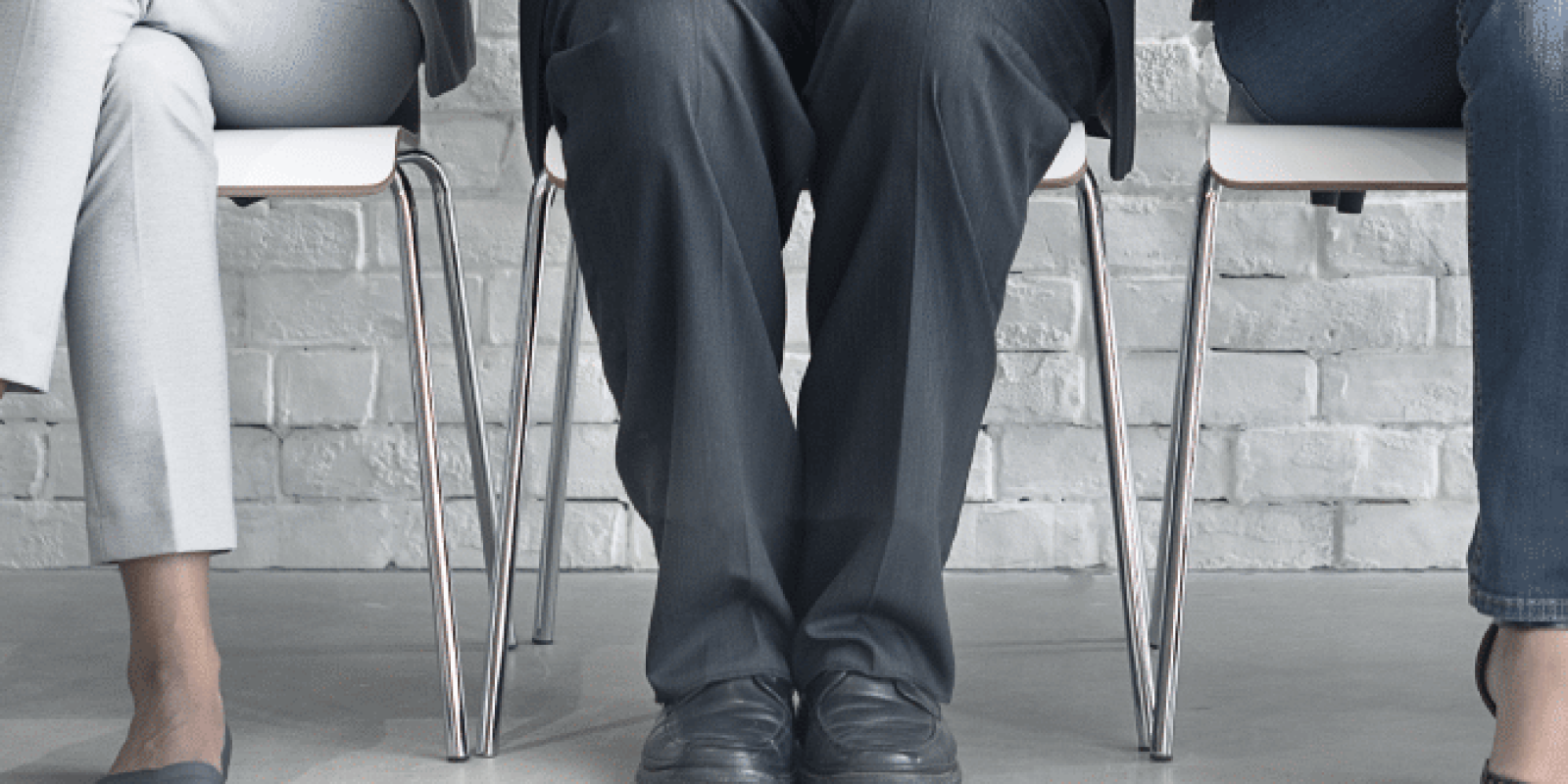 Three people in business attire sit on chairs against a white brick wall, each holding a smartphone or tablet, with only their lower bodies visible.