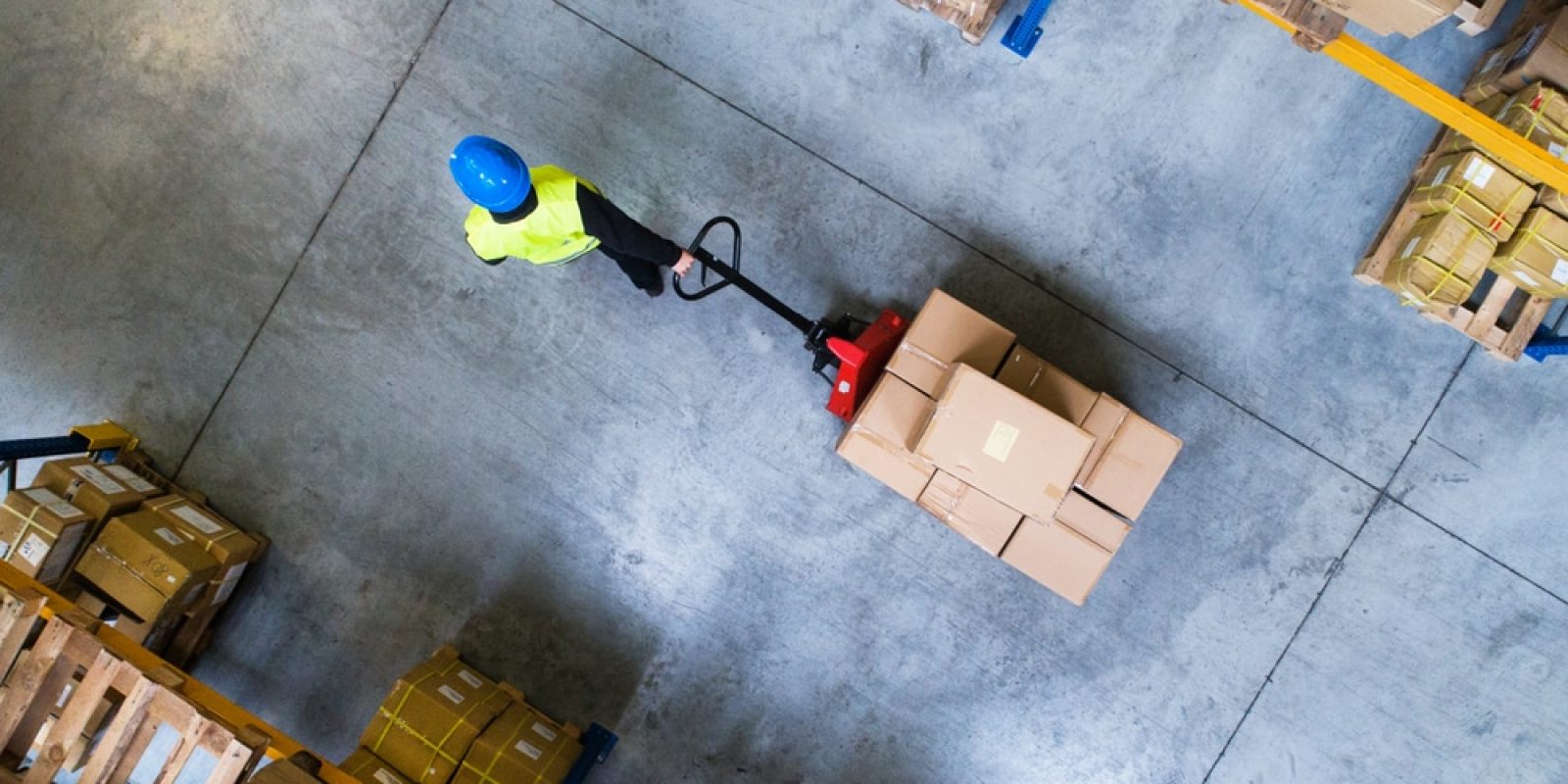 A warehouse worker wearing a blue helmet and yellow vest moves a pallet of boxes with a manual pallet jack, surrounded by stacks of cardboard boxes on pallets, viewed from above.