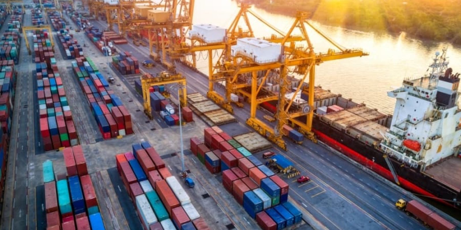 Aerial view of a busy 3PL shipping port with large yellow cranes, stacked colorful shipping containers, and a cargo ship docked by the water at sunset.