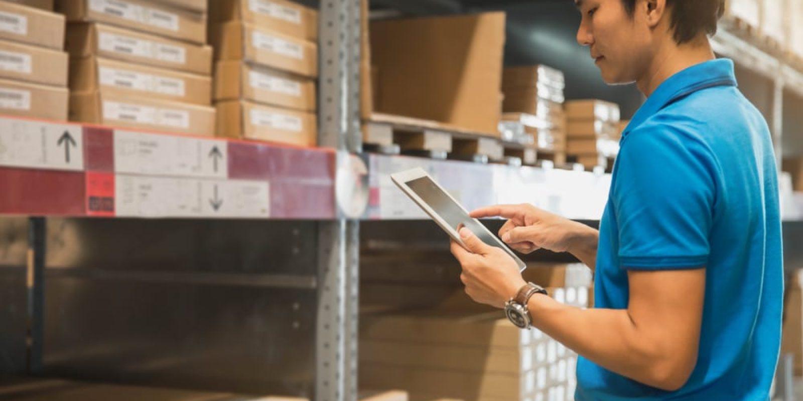 A man in a blue polo shirt uses a tablet while standing in a warehouse aisle of micro-fulfillment centers, surrounded by shelves stacked with labeled cardboard boxes.
