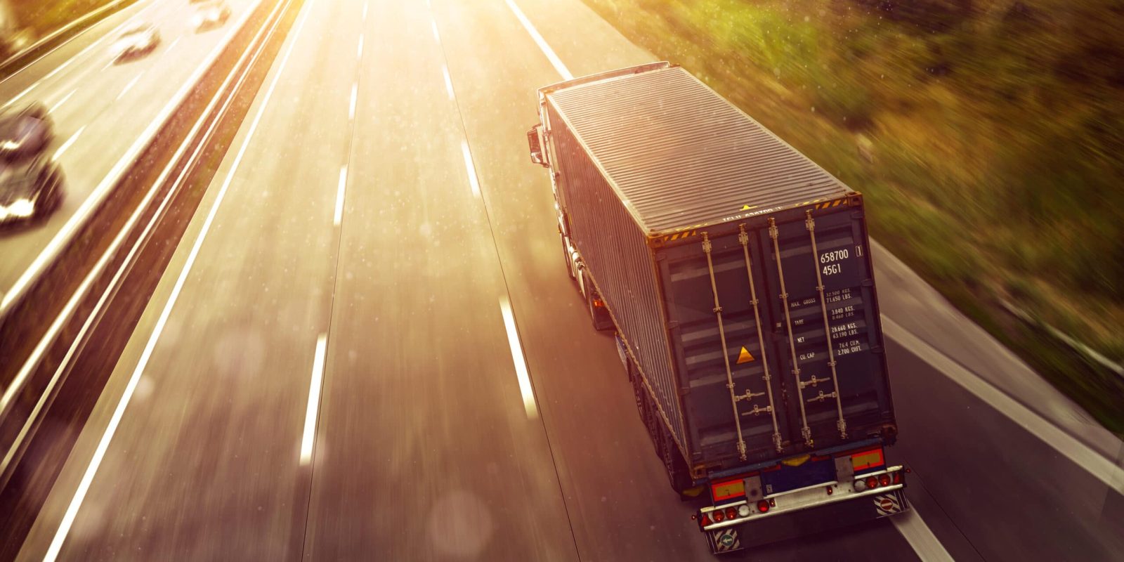 A container truck drives on a sunlit highway, surrounded by greenery, with a few cars visible in other lanes. The bright sunlight creates a warm, glowing effect over the road.