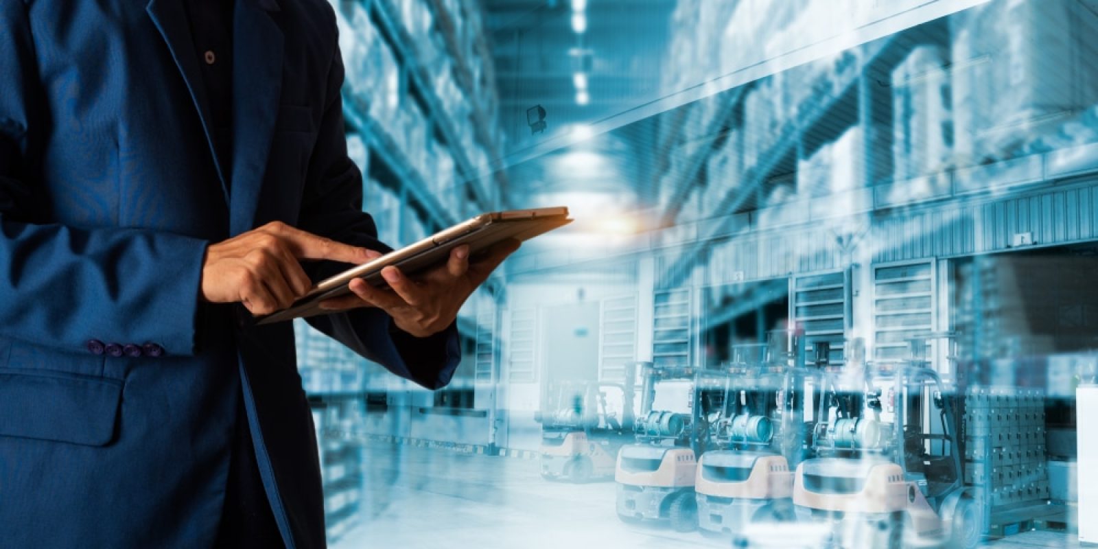 A person in a blue suit uses a tablet, with a modern warehouse filled with shelves and forklifts in the background, symbolizing logistics, inventory management, or supply chain planning operations.