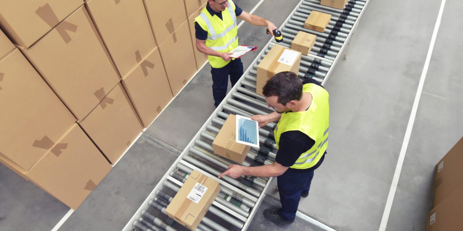 Two workforce members in yellow vests scan and check boxes on a conveyor belt in a warehouse. Stacks of cardboard boxes are visible nearby, and both workers are handling packages.