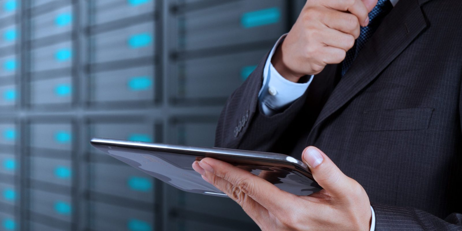 A man in a suit adjusts his tie while holding a tablet, standing in front of rows of server racks in a data center, highlighting the efficiency of goods-to-person technology in modern logistics operations.