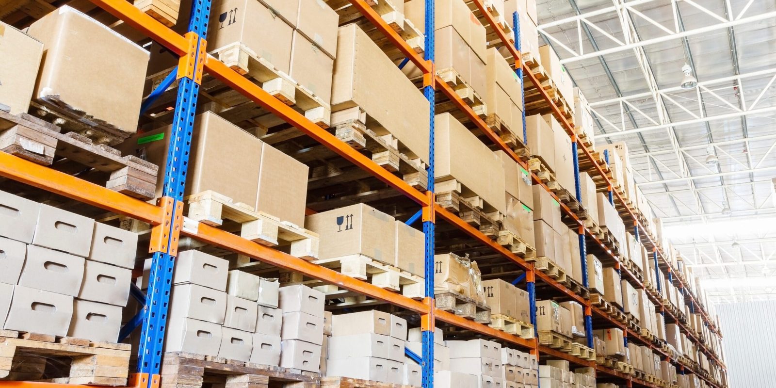 Rows of tall warehouse shelves filled with stacked cardboard boxes and packages, organized on wooden pallets in a brightly lit industrial storage facility.