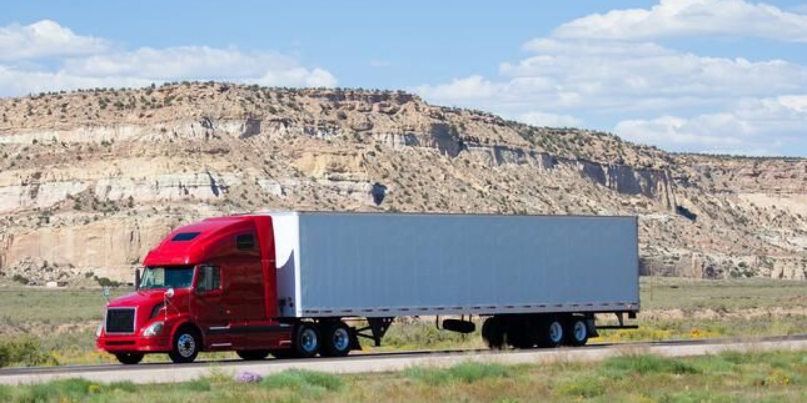 A red semi-truck with a white and gray trailer drives along a highway, with rocky, tan cliffs and a blue sky with scattered clouds in the background.