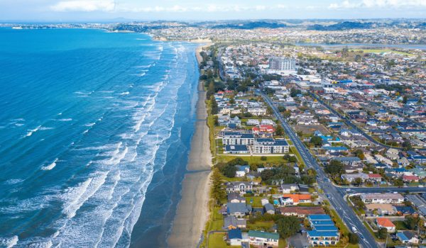 Aerial view of a coastal city with houses and buildings along a sandy beach. Waves from the blue ocean break along the shore, and a road runs parallel to the coastline in this stunning envista. The city extends inland with scattered greenery.