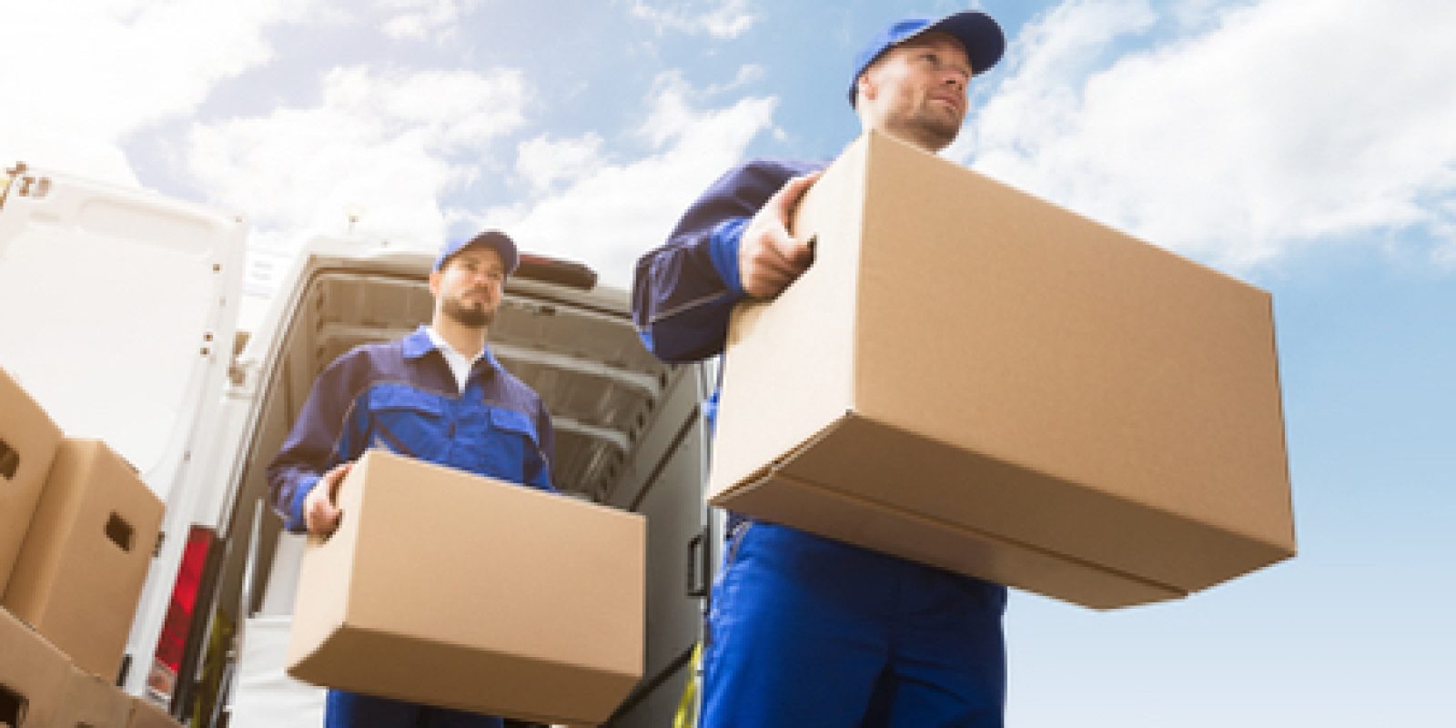 Two movers wearing blue uniforms carry cardboard boxes from the back of a white van under a bright blue sky with scattered clouds. More boxes are stacked nearby.
