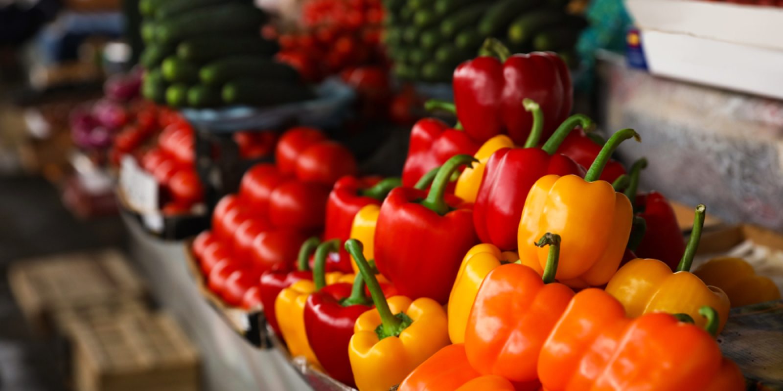 Colorful bell peppers—orange, yellow, and red—are displayed in rows at a market stall, with cucumbers and cherry tomatoes visible in the background, offering a vibrant scene reminiscent of cloud computing’s seamless organization.