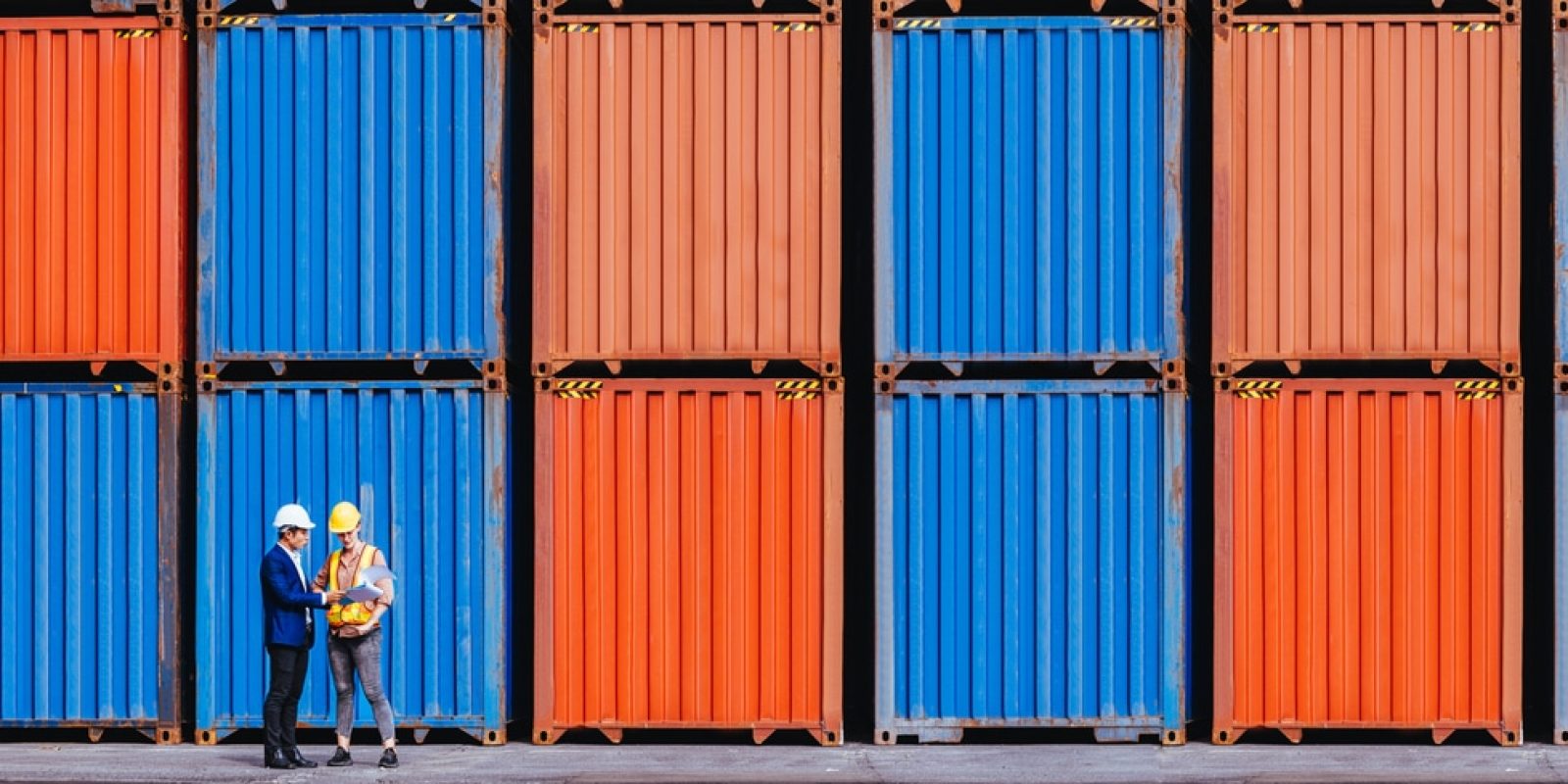 Two people wearing helmets stand in front of stacked orange and blue shipping containers, appearing to have a discussion at a shipping yard or port.