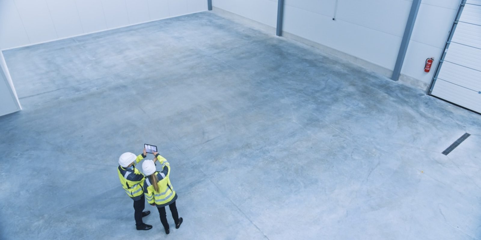 Two construction workers in high-visibility jackets and hard hats stand in an empty, spacious industrial warehouse, looking at a tablet together. The warehouse has a concrete floor and white walls.
