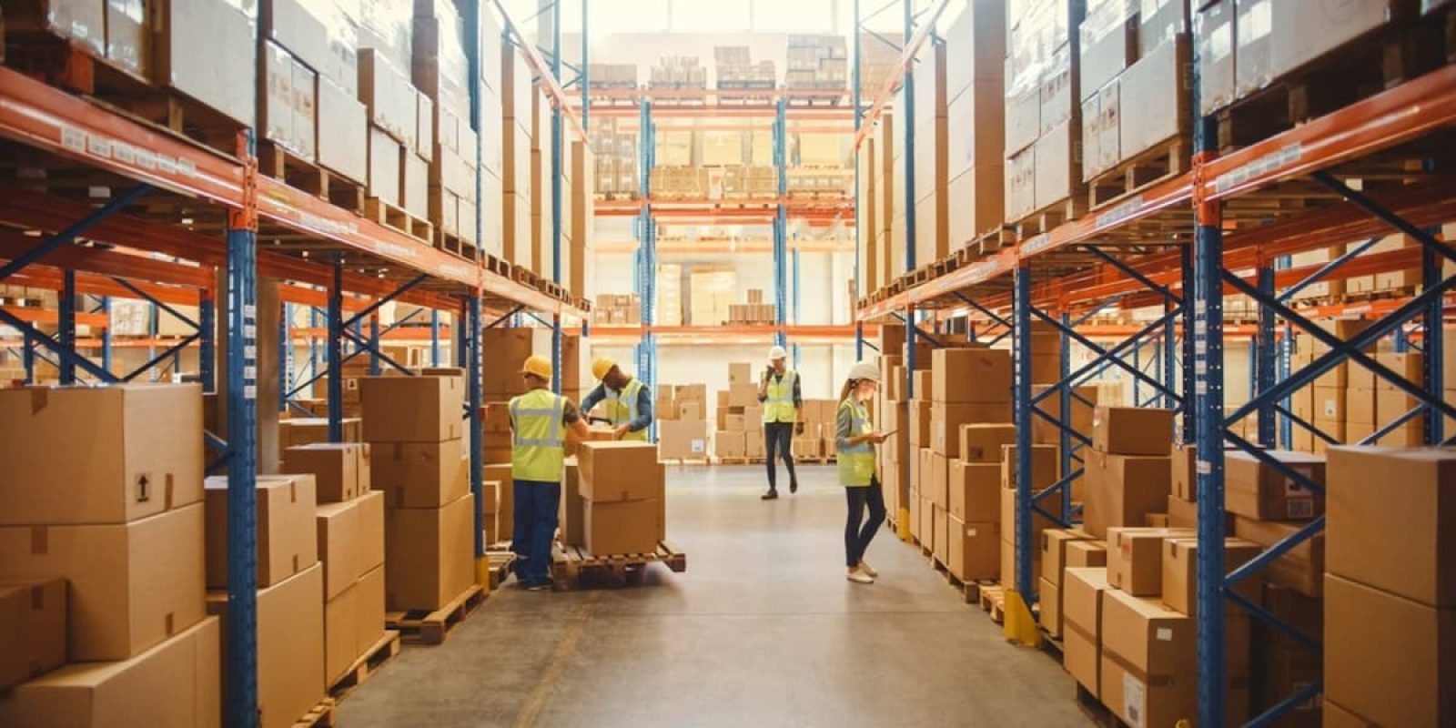 Workers wearing safety vests organize and move cardboard boxes in a large, well-lit warehouse filled with shelves stacked with more boxes.