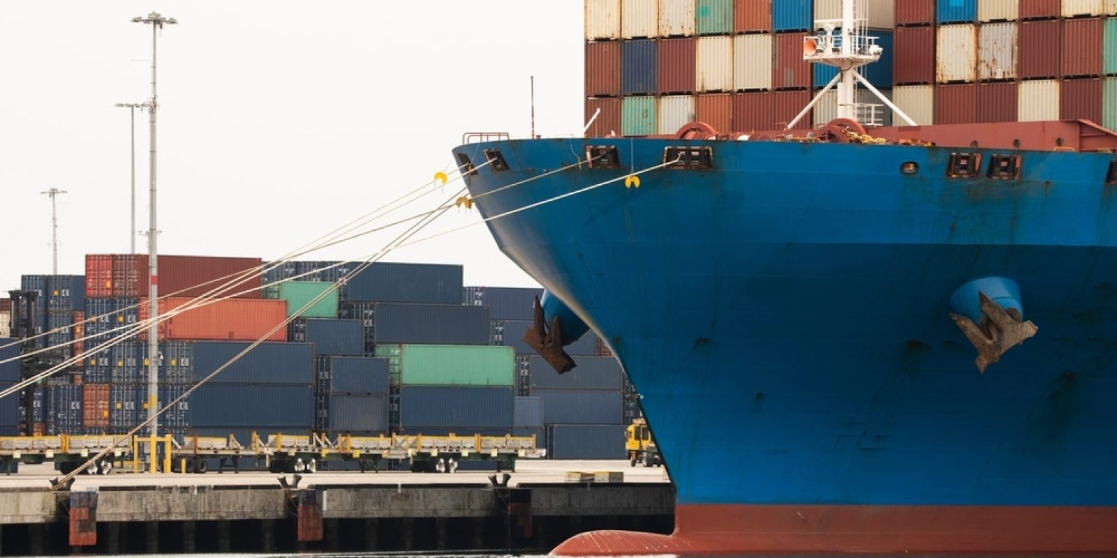 A large blue cargo ship docked at a port, with colorful shipping containers stacked both on the vessel and on the dock in the background.