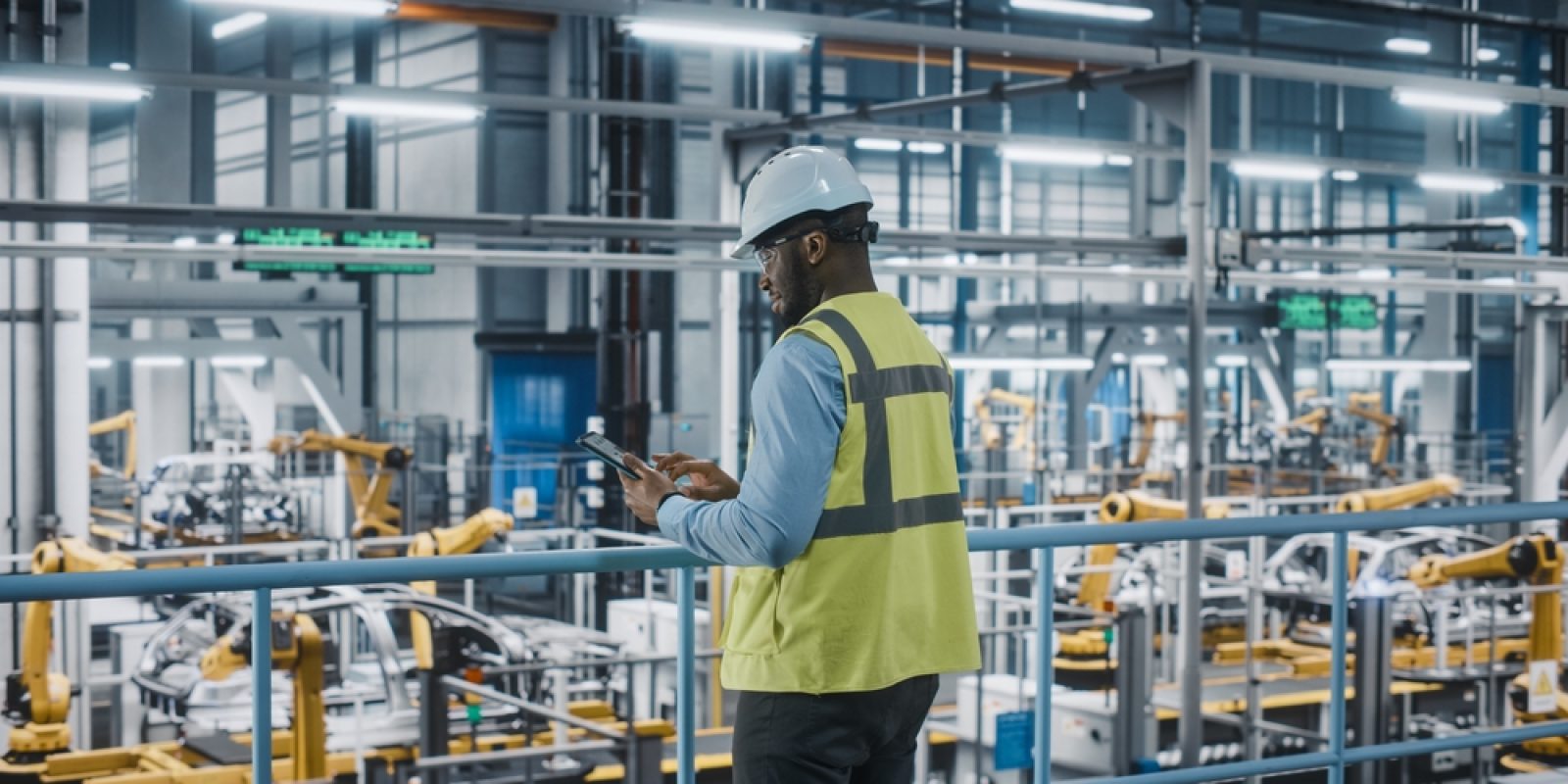 A person wearing a white hard hat and yellow safety vest uses a tablet while standing in a modern, automated factory with robotic arms and machinery in the background.