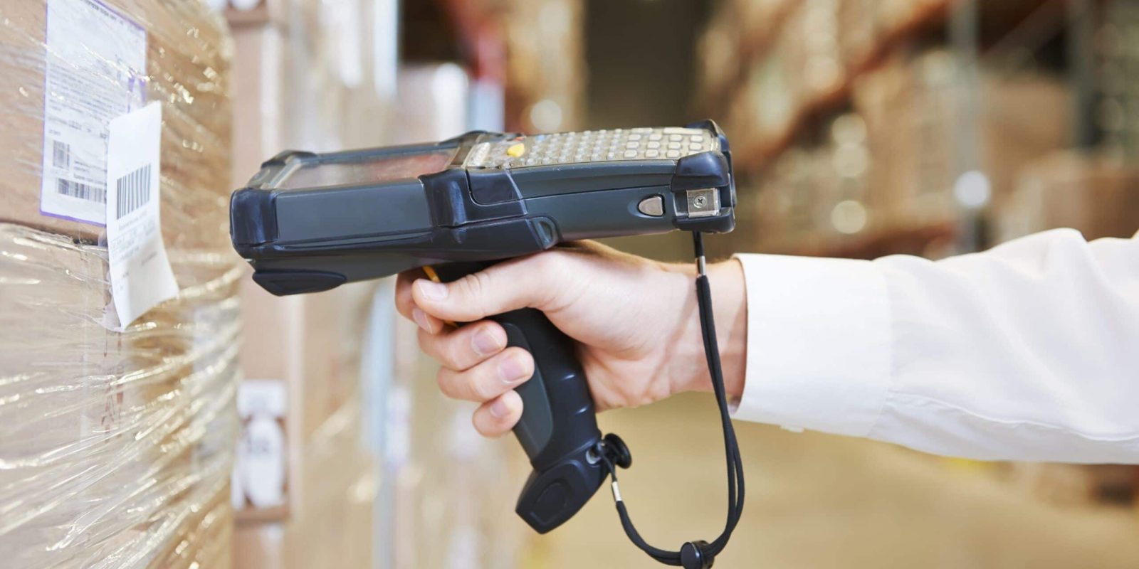 A person scans a barcode on a pallet of boxes in a warehouse using a handheld barcode scanner, helping ensure food traceability. Shelves filled with products are visible in the background.