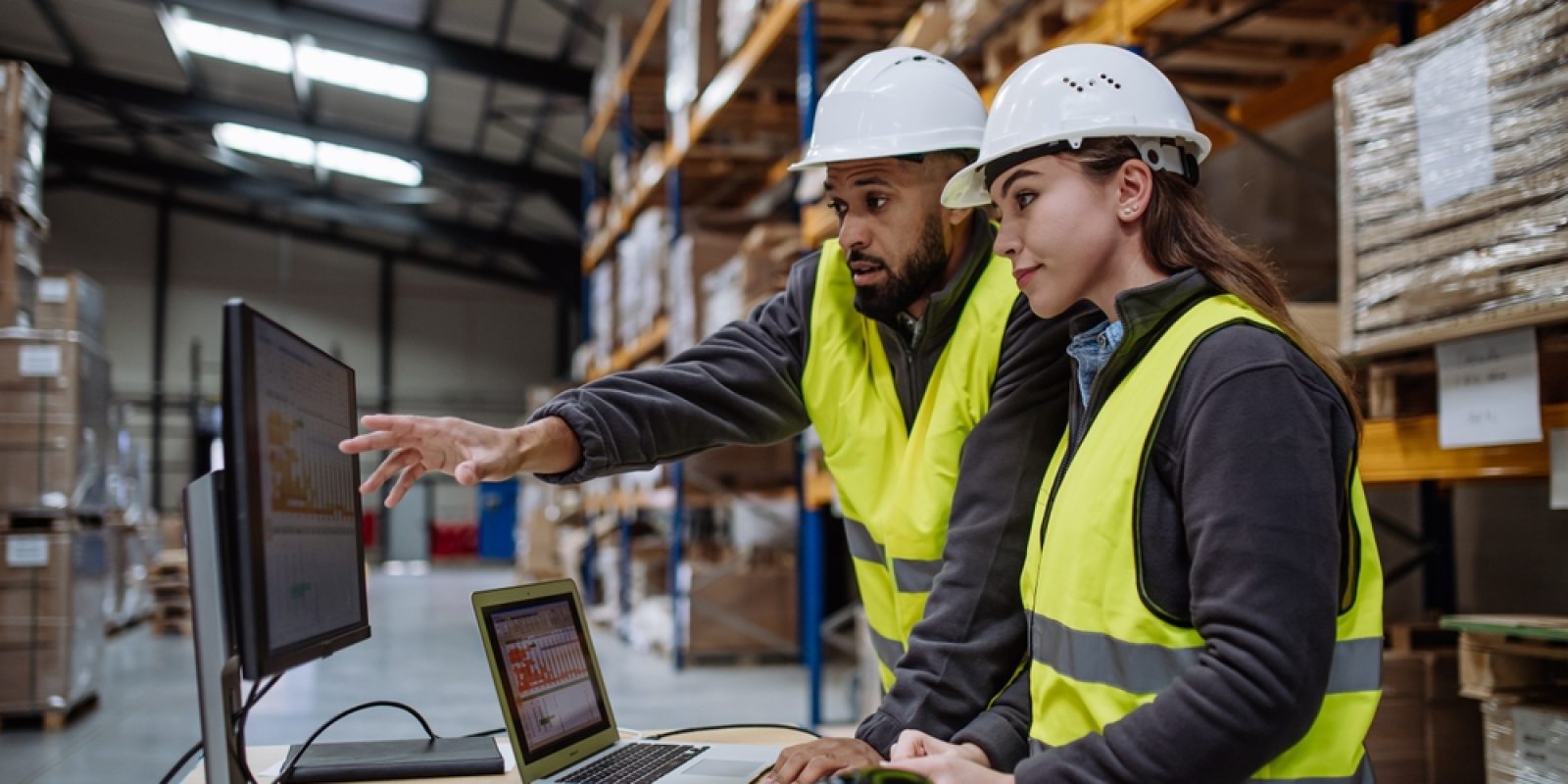 Two warehouse workers in safety vests and hard hats review order accuracy on a computer monitor in a storage facility, surrounded by shelves with stacked boxes and paperwork on a desk.