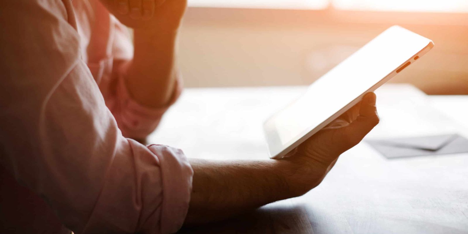 A person in a light pink shirt sits at a table, holding a digital tablet in one hand and resting their chin on the other hand, with soft sunlight streaming in through a window in the background.