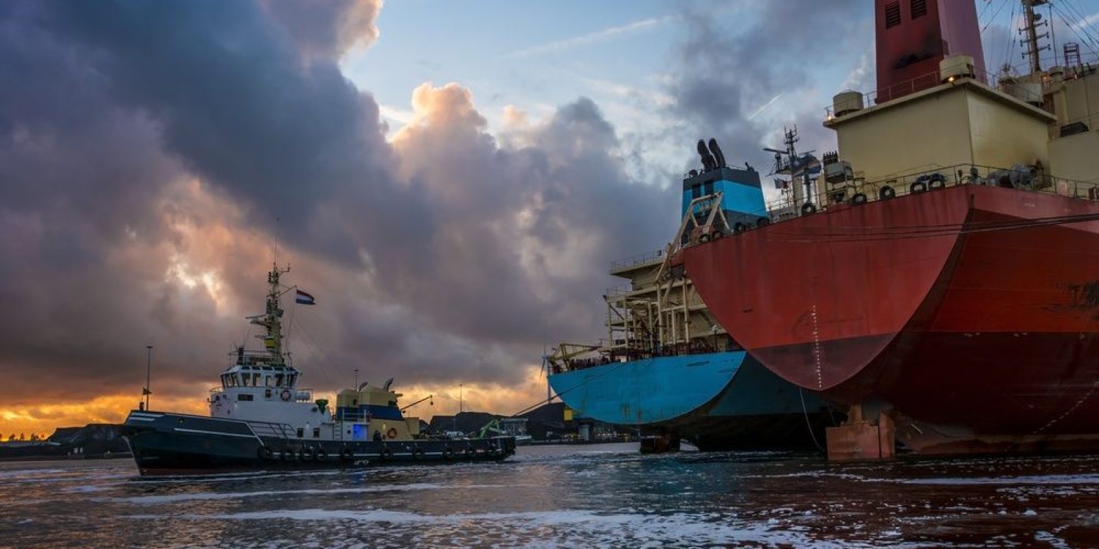 A tugboat moves near two large docked cargo ships at sunset, with dramatic clouds and golden light reflected on the water.