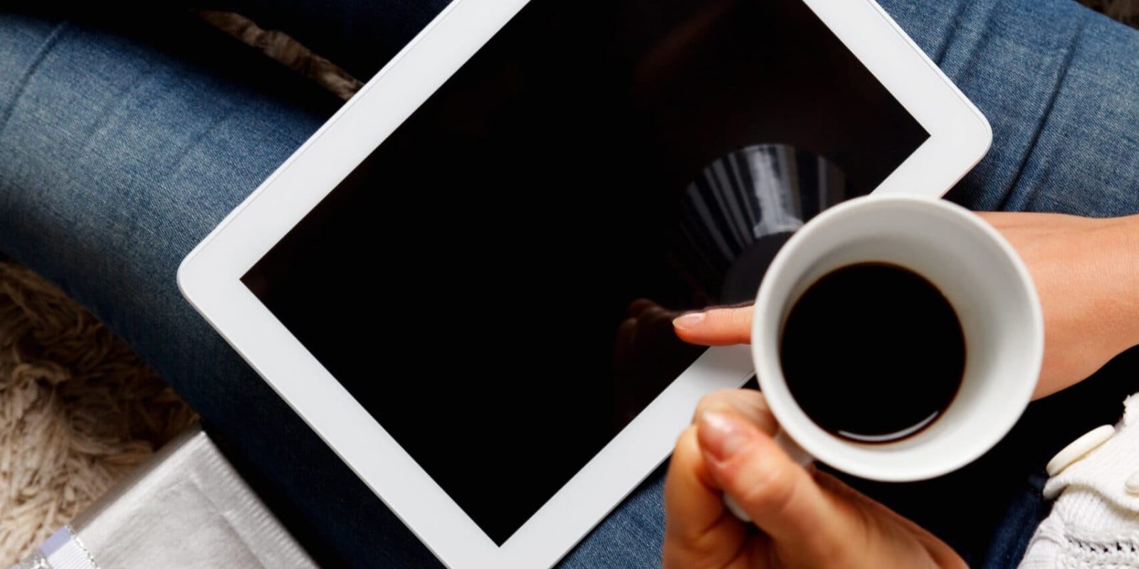 A person sitting with wrapped gifts holds a cup of coffee and a tablet, with a credit card nearby, suggesting online holiday shopping or gifting.