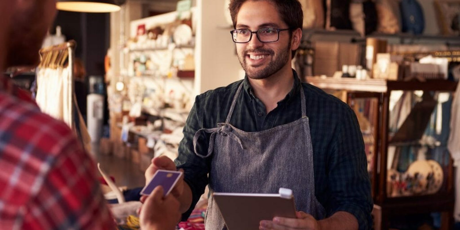 A smiling shop worker in an apron holds a tablet and processes a payment from a customer handing over a credit card in a cozy store environment. Shelves with various items are visible in the background.
