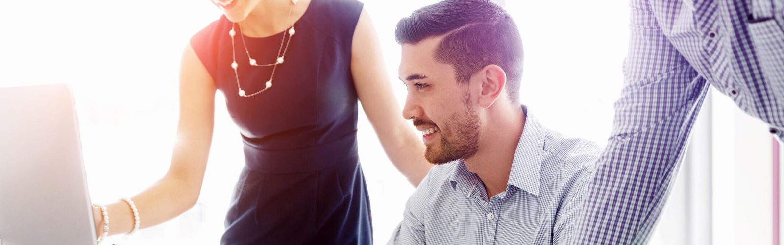 Three colleagues smile and collaborate at a desk, looking at a computer screen. One woman stands pointing at the monitor, while two men sit and stand nearby, engaged in discussion in a bright office setting.