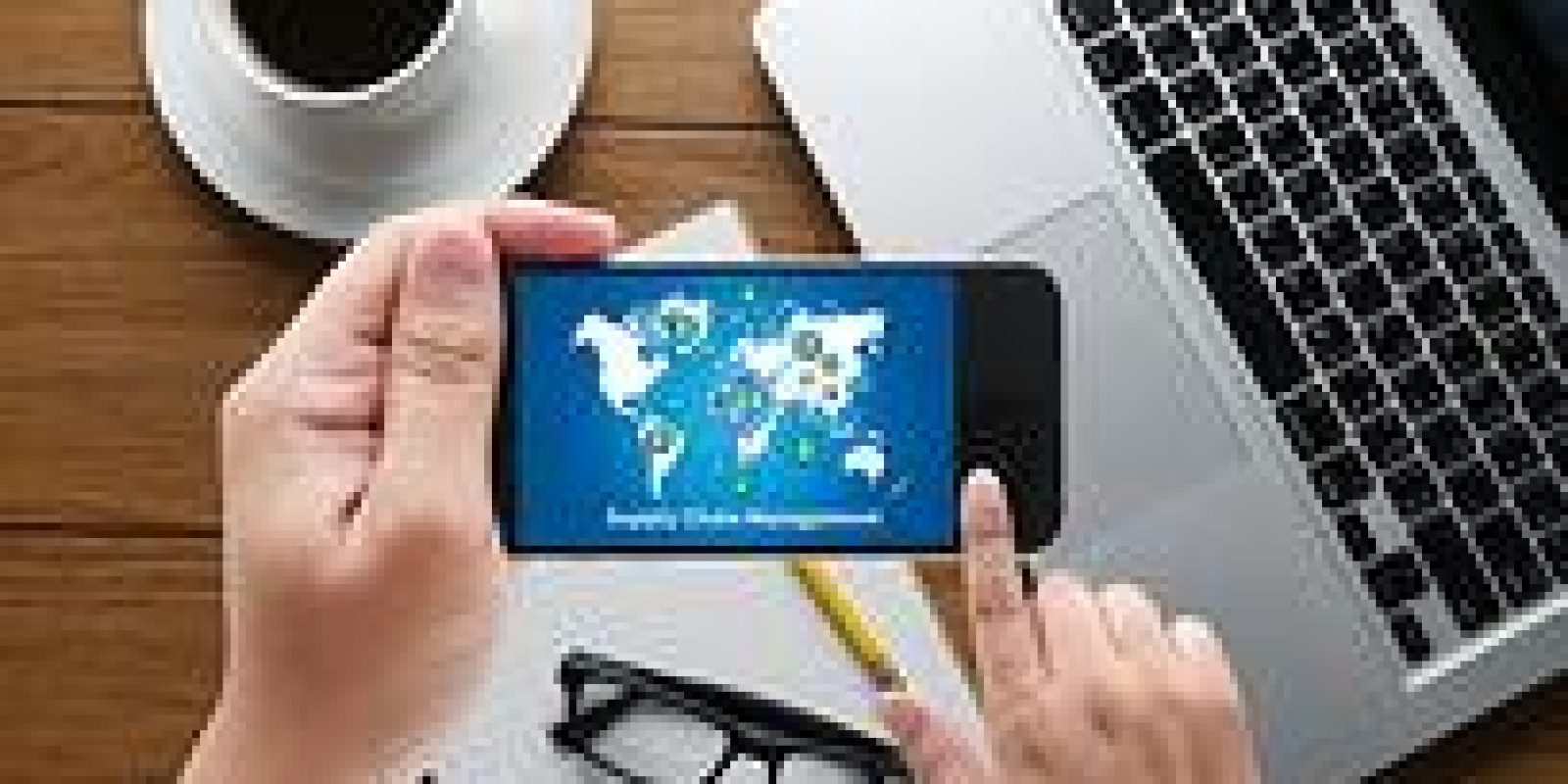 A person holds a smartphone displaying a world map with network icons. The phone is above a notepad, eyeglasses, and a keyboard, with a cup of coffee nearby on a wooden desk.