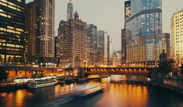Downtown cityscape at dusk with illuminated envista skyscrapers, a lit-up bridge over the river, and blurred boats gliding across the water, reflecting city lights.