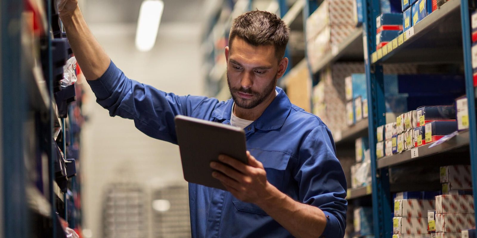 A man in a blue work jumpsuit stands between warehouse shelves, reaching for a box with one hand and holding a tablet in the other, checking inventory or stock.