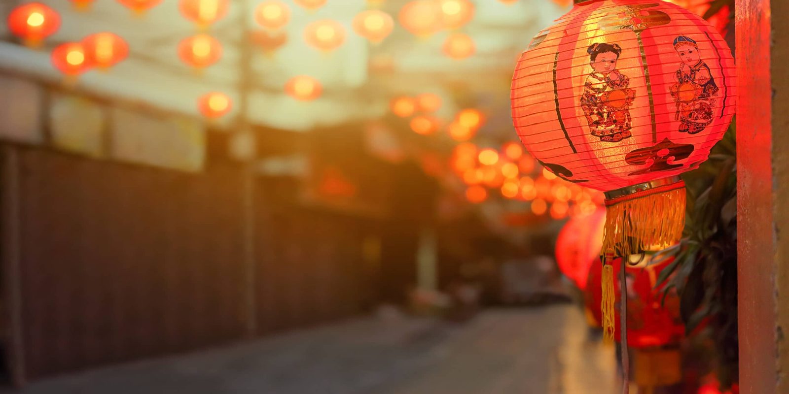 A red paper lantern with an illustration of a woman in traditional attire hangs outside, glowing among many similar lanterns lining a street in the evening. The background is softly blurred, emphasizing the lanterns’ warm light.