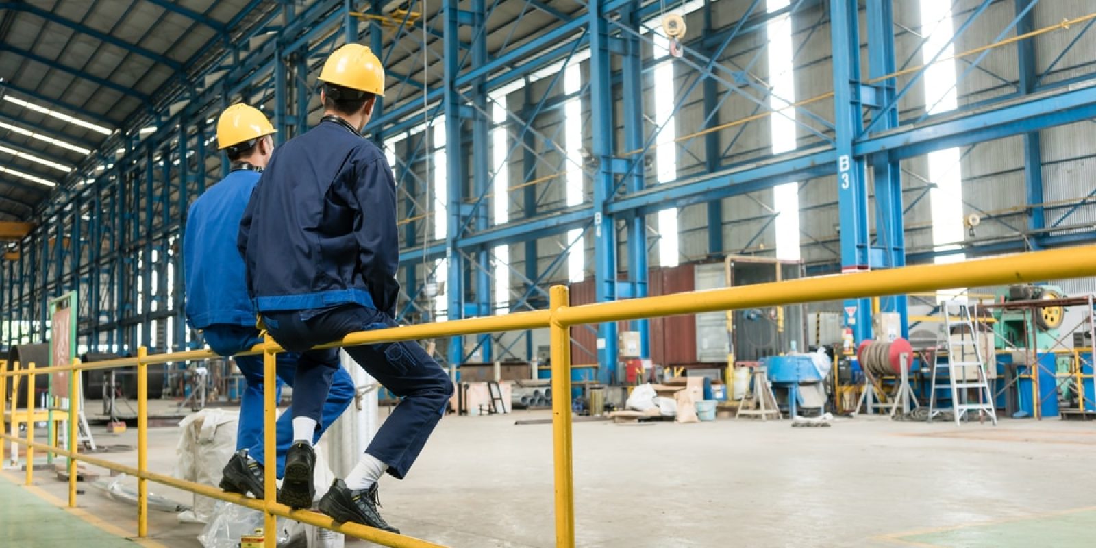 Two workers in blue uniforms and yellow hard hats sit on a yellow metal railing inside a spacious industrial warehouse with high ceilings and blue structural beams.
