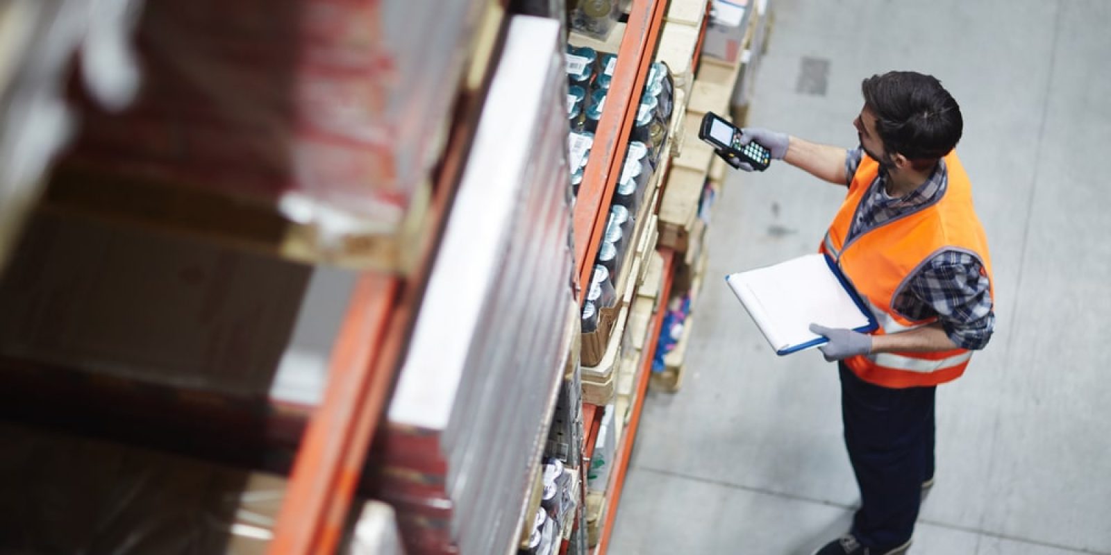 A warehouse worker in an orange safety vest scans items on a high shelf with a handheld device while holding a clipboard, standing in an industrial storage area.