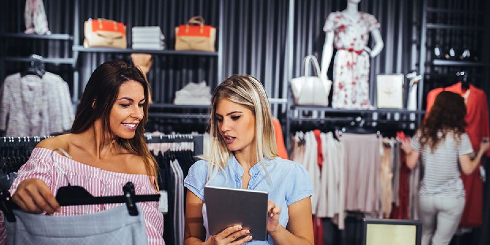 Two women stand in a clothing store, one holding a skirt and the other using a tablet. Clothing racks and accessories are visible, with another person browsing in the background. Mannequins display outfits.
